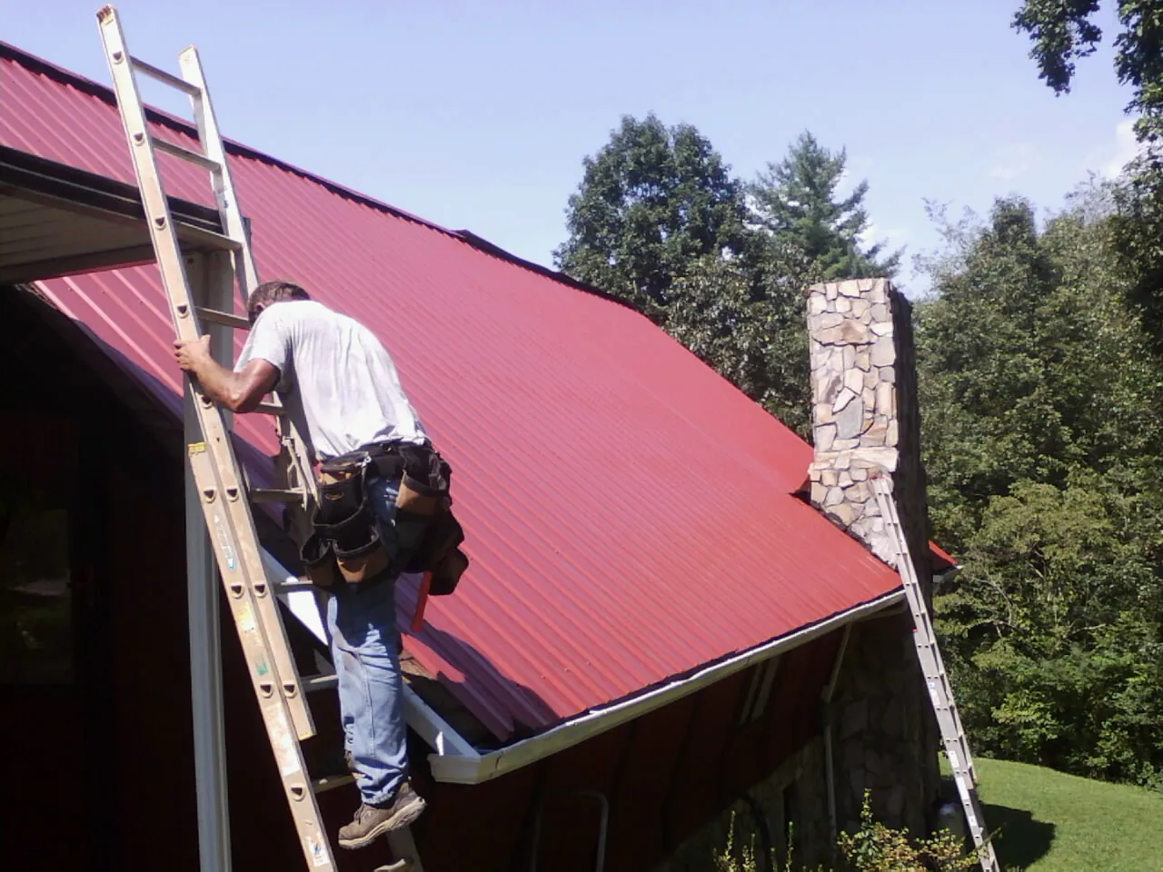 Roofer on a ladder beside a red metal roof and stone chimney — Soffit & Fascia Repair by Roofing Brothers in Brackenridge
